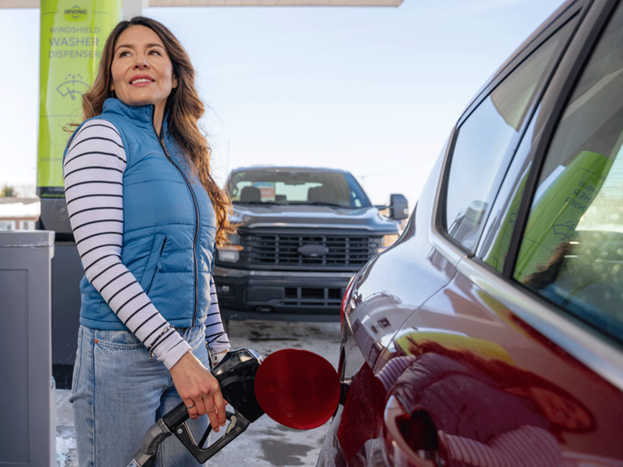 Person in a blue vest holds a fuel nozzle to a red car at a gas pump; a green windshield washer station and another vehicle are visible in the background.